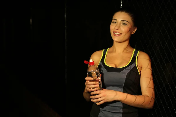Menina fitness jovem com garrafa de água no fundo escuro — Fotografia de Stock