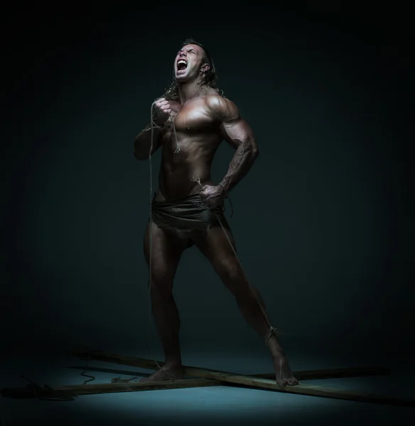 Screaming naked muscular man, hands tied rope to wooden beams