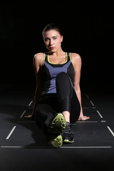 Cute young fitness woman sitting on floor over black background — Stockfoto