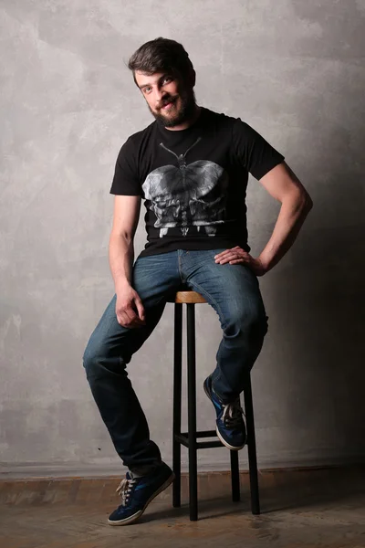 Homme barbu en T-shirt noir avec assise sur un tabouret de bar. Gris — Photo