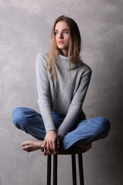 Menina sentada em suéter com pernas cruzadas. Fundo cinzento — Fotografia de Stock