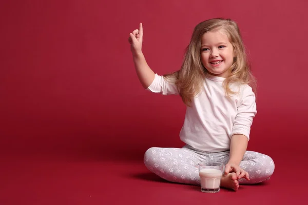 Baby girl in pajamas with glass of milk showing finger. Red background