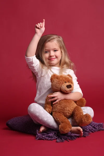 Flicka i pyjamas med en björn som visar ett finger. Röd bakgrund — Stockfoto