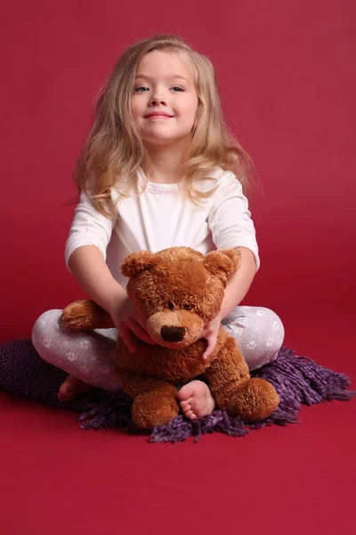 Liten flicka i pyjamas Holding Bear. Röd bakgrund — Stockfoto