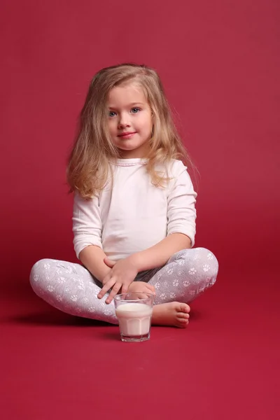 Sittande bebis i pyjamas med glas mjölk. Röd bakgrund — Stockfoto