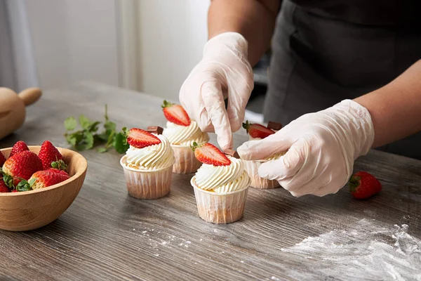 Closeup of a pastry chef decorating cupcakes with fresh strawberries and chocolate, on a table.