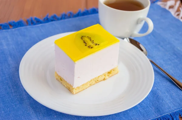 Delicado pastel de postre de cuajada con kiwi de gelatina en un plato blanco en una servilleta azul y una taza de té . — Foto de Stock