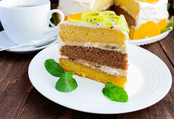 Un morceau de gâteau aux fruits (kiwi, orange, feuilles de menthe) sur une assiette blanche sur fond de bois — Photo