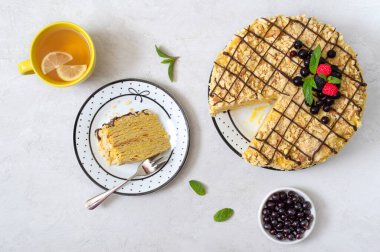 Böğürtlenlerle süslenmiş çok katmanlı vanilyalı pasta, nane. Tabakta bir parça lezzetli kek ve limonlu bir fincan sabah çayı. Üst görünüm.