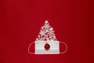 Kırmızı Noel mücevheri ve kar taneleri kırmızı ve koyu renk bir arka planda beyaz cerrahi maskeli bir noel ağacı şeklinde serilmiştir.