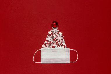 Kırmızı noel balosu ve kar taneleri kırmızı ve koyu renk bir arka planda beyaz cerrahi maskeli bir noel ağacı şeklinde serilmiştir.