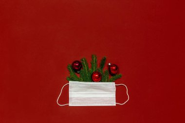 Kırmızı noel baloları ve yeşil noel ağacı dalları. Kırmızı monoponik arka planda beyaz tıbbi maske.