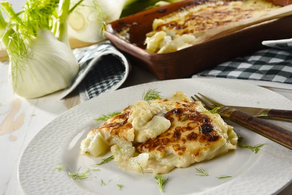 Pişmiş rezene au gratin beyaz bir tabakta servis ediliyor, yanında da taze rezene var. 