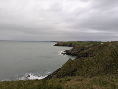 Saf yeşil kayalıklar İrlanda 'nın Waterford ilçesindeki Boatstrand yakınlarındaki Copper Coast boyunca uzanan sakin kıyı şeridine doğru iniyor. Sönük ışık, bulutlu gökyüzü ve uzak burunlar engebeli arazi ve suyla işaretlenmiş sakin ama dramatik bir kıyı manzarası yaratır.