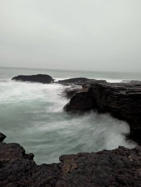 Wexford County 'deki Kanca Kafa yakınlarındaki Kelt Denizi' ne güçlü dalgalar çarptığında kıyıdan karanlık ve açısal kaya oluşumları yükseliyor. Bulutlu gökyüzü ve çalkantılı su, Ireland 'ların engebeli kıyı manzarasını dramatik ve atmosferik bir şekilde gösteriyor.