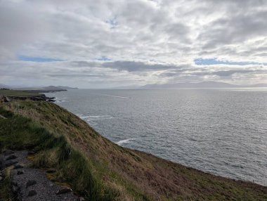 The cliffs along Slea Head Drive rise above the Atlantic with a sweeping curve of coastline leading toward distant headlands. Soft, filtered light breaks through the cloud cover and reflects on the water, while the grassy cliff edge in the foreground