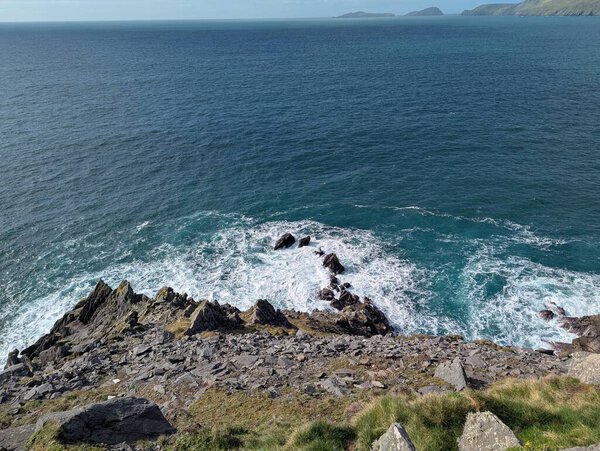 Rocky cliffs drop sharply into the Atlantic Ocean along Slea Head Drive, where waves crash against the rugged shoreline beneath sweeping views of distant headlands. The scene captures the dramatic natural beauty of Irelands western coast