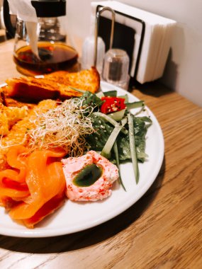 Sıcak bir kafe ortamında füme somon, salata ve kızarmış ekmek ile renkli kahvaltı tabağı.