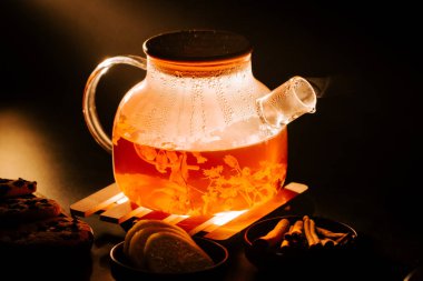 Black tea brewing in a glass teapot with cookies and spices on a dark table
