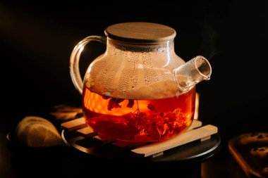 Steam rises from a pot of black tea with flowers on a wooden stand at evening time