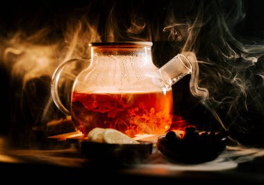 Steam rises from a glass teapot filled with black tea and lemon on a dark surface