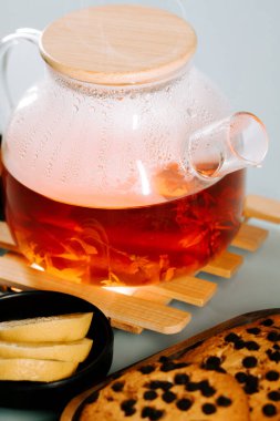 Warm tea steeping in a glass pot with cookies and lemon slices on a wooden surface