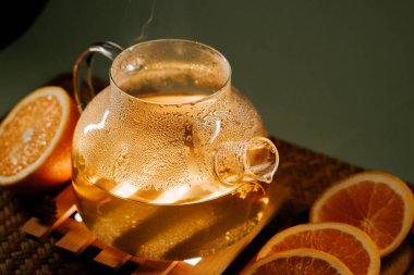 Steaming green tea in a glass teapot with fresh orange slices on a wooden tray