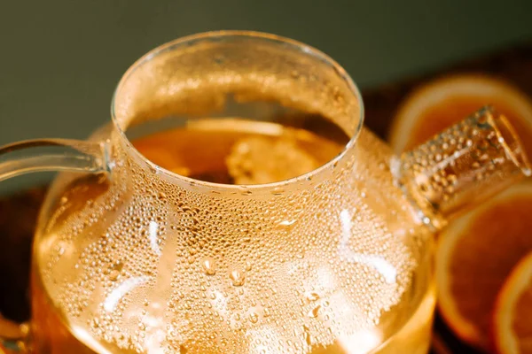Steam rises from a glass teapot filled with green tea beside fresh orange slices