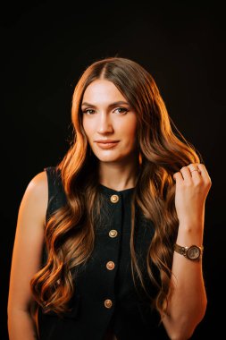 Uzun saçlı bir kadın, zarif bir elbise içindeki koyu bir arka plana karşı özgüvenle poz verir.