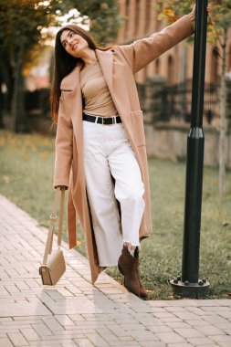 Bej ceketli şık bir kadın güneşli bir sonbahar öğleden sonrasında parkta geziniyor.