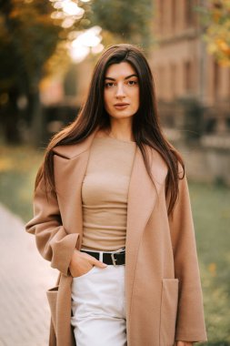 Şehirde huzurlu bir sonbahar yolu boyunca şık bir mantoyla poz verir.