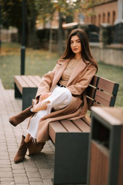 Parkta bankta oturan genç bir kadın sonbaharda şık bir kıyafet giyiyor.