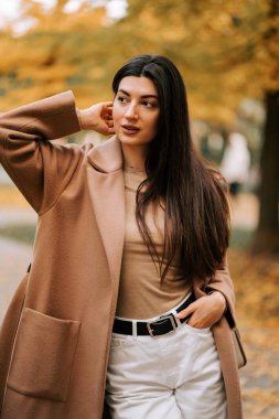 Gündüz vakti sonbahar renkleriyle dolu bir parkta poz veren şık ceketli bir kadın.