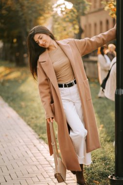 Parkta güneşli bir günün tadını çıkaran şık ceketli bir kadın sokak lambasının yanında poz veriyor.