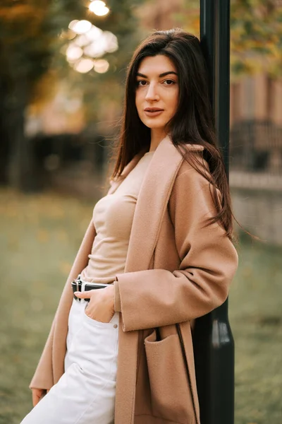 Sonbaharda bir parkta direğe yaslanmış şık kıyafetli genç bir kadın.