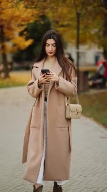 Sonbahar öğleden sonra telefonuna odaklanmış genç bir kadın parkta yürüyor.