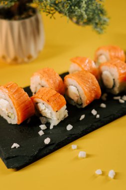 Lezzetli somonlu suşi dürümleri sarı arkaplanlı siyah bir tabakta servis ediliyor.