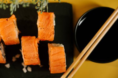 Siyah tabakta servis edilen ızgara somonlu taze suşi ruloları ve sarı arka plan.