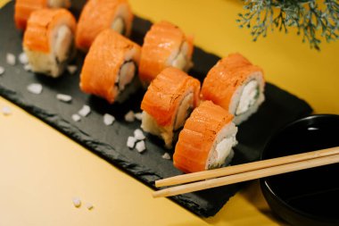 Sarı arka planda çubuklarla siyah bir tahta üzerinde renkli somon suşisi yuvarlanıyor.