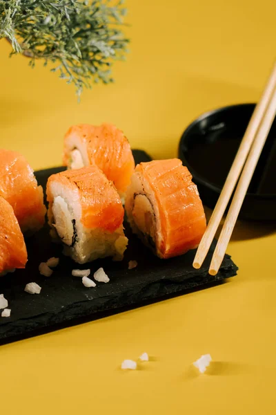 Lezzetli suşi dürümleri, siyah bir tabakta, canlı sarı bir arka planda yemek çubukları ile.