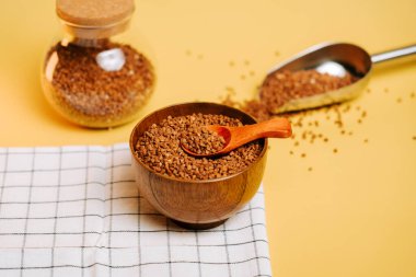 Karabuğday, cam kavanozun yanında ahşap bir kasede inliyor ve sarı arka planda kepçeyle...