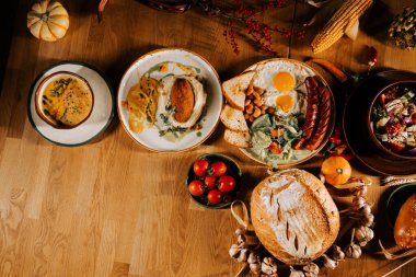 Kahvaltı, yumurta, sosis, ekmek, çorba ve taze sebzelerle ahşap bir masada.