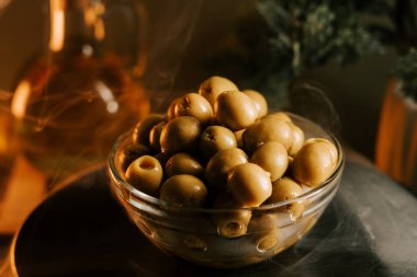 Temiz cam bir kasede servis edilen taze yeşil zeytinler ve sıcak bir arka plan.