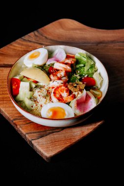 Tahta tabakta servis edilen karides, yumurta ve taze sebzeli renkli salata kasesi.