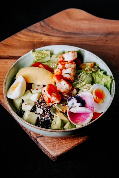 Tahta bir tahtanın üzerinde çeşitli sebzeler, proteinler ve yenilebilir çiçeklerle servis edilen sağlıklı salata kasesi.