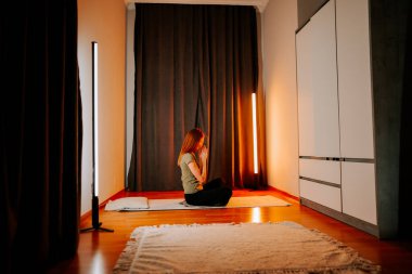 Genç bir kadın akşam saatlerinde evinde hafif ışıklarla yoga yapıyor.