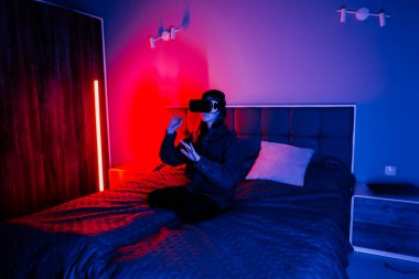 Genç bir kadın, yatakta otururken mavi ve kırmızı ışıklı bir odada sanal gerçekliği keşfediyor.