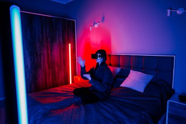 Genç bir kadın akşam saatlerinde renkli ışıklarla yatak odasında sanal gerçekliği deneyimliyor.