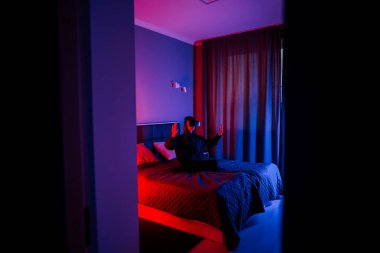 Genç bir kadın, kapalı bir odada yatakta otururken sanal gerçeklik gözlüğü kullanıyor.