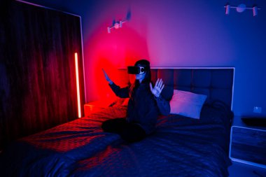 Genç bir kadın karanlık bir odada yatağında otururken sanal gerçekliği deneyimliyor.